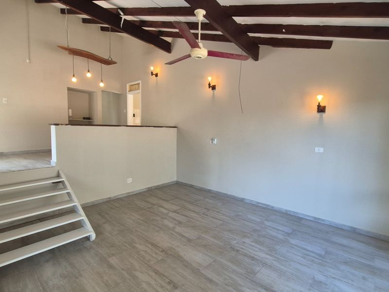 An open-plan living area featuring exposed timber ceiling beams, tiled flooring, and a split-level layout with a staircase and wall-mounted lighting.