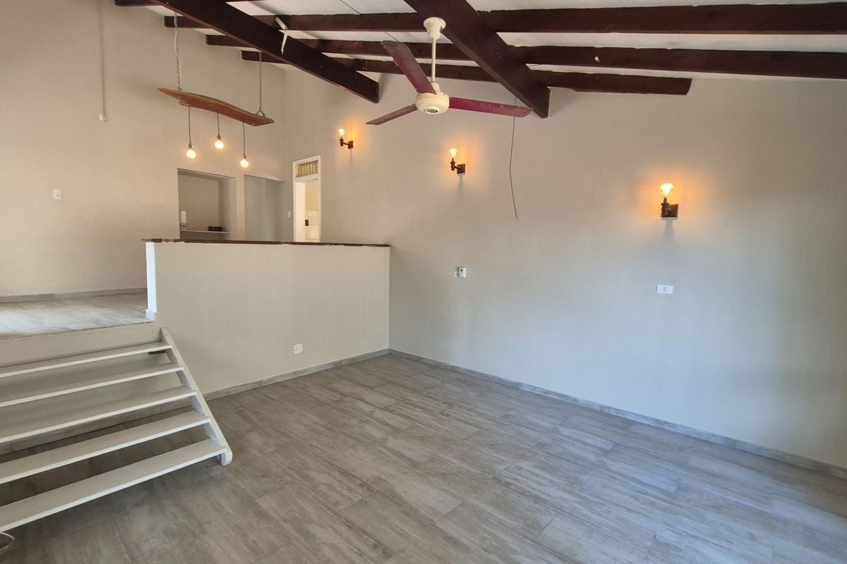 An open-plan living area featuring exposed timber ceiling beams, tiled flooring, and a split-level layout with a staircase and wall-mounted lighting.