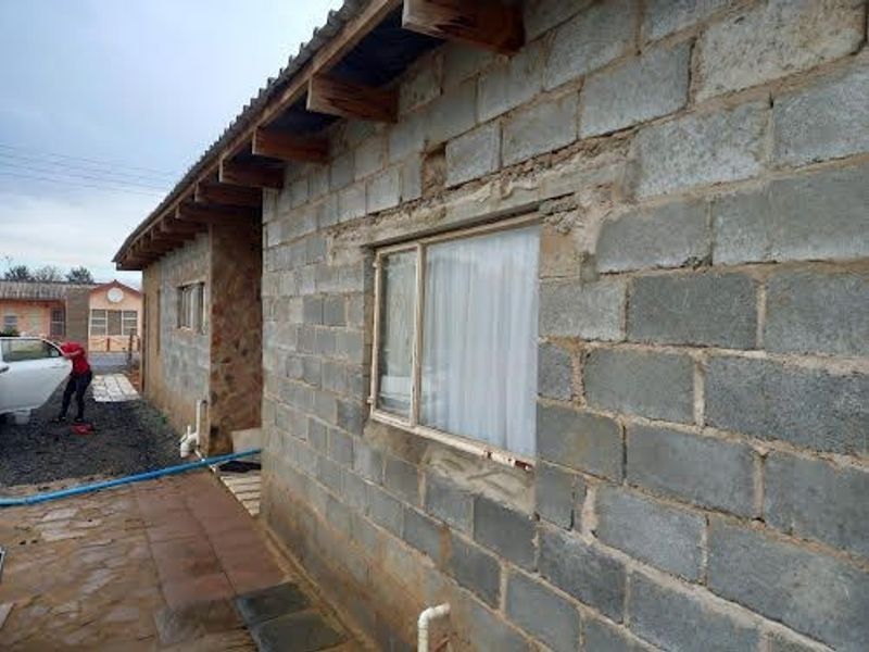 Exterior view of a house featuring exposed concrete block walls, timber roof rafters, and stone cladding, currently in an unfinished state.