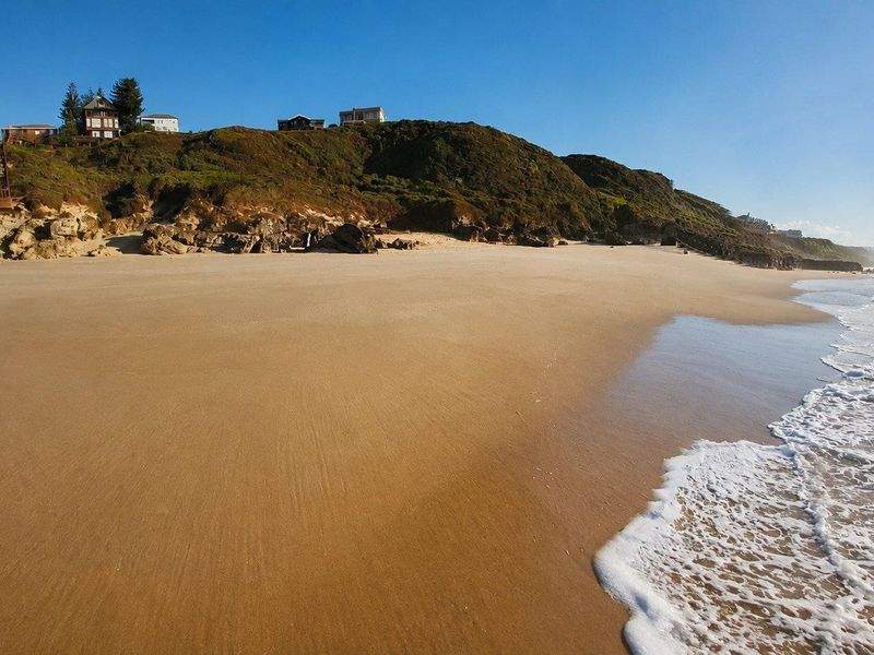 A scenic view of a sandy beach with waves crashing on the shore, featuring residential properties situated on the clifftop above.