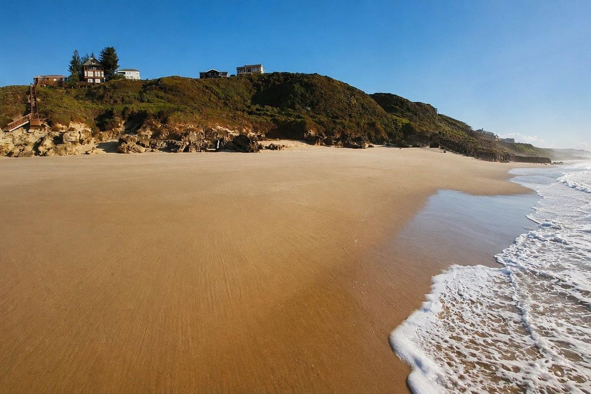 A scenic view of a sandy beach with waves crashing on the shore, featuring residential properties situated on the clifftop above.