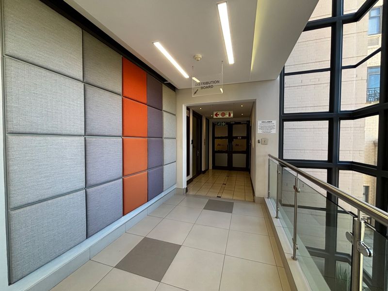 A modern, well-maintained office hallway featuring acoustic wall paneling, tiled flooring, recessed linear lighting, and a glass balustrade overlooking the exterior.