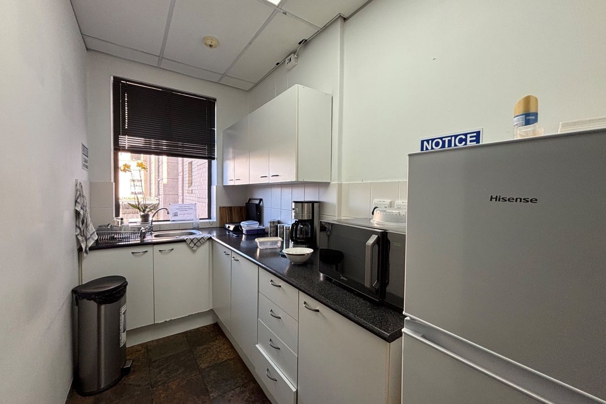 A functional office kitchenette featuring white built-in cabinetry, dark laminate countertops, a stainless steel sink, and durable tiled flooring.