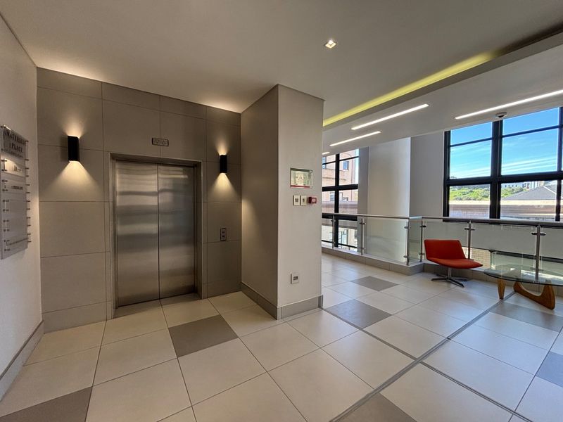 Modern office building lobby featuring a stainless steel elevator, large windows with natural light, and high-quality tiled flooring.