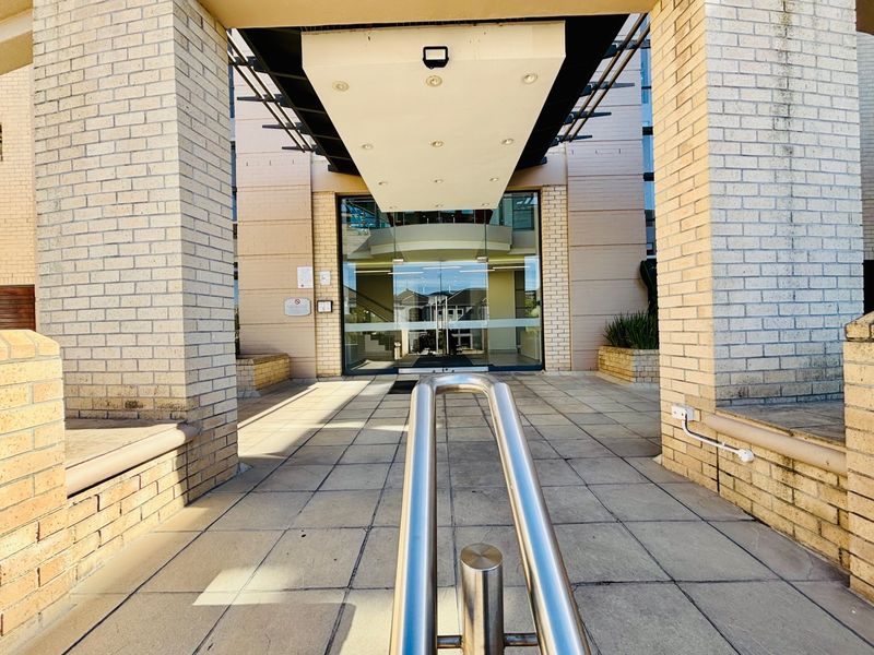 Main office building entrance featuring a paved walkway, stainless steel handrail, and a covered canopy leading to glass double doors.