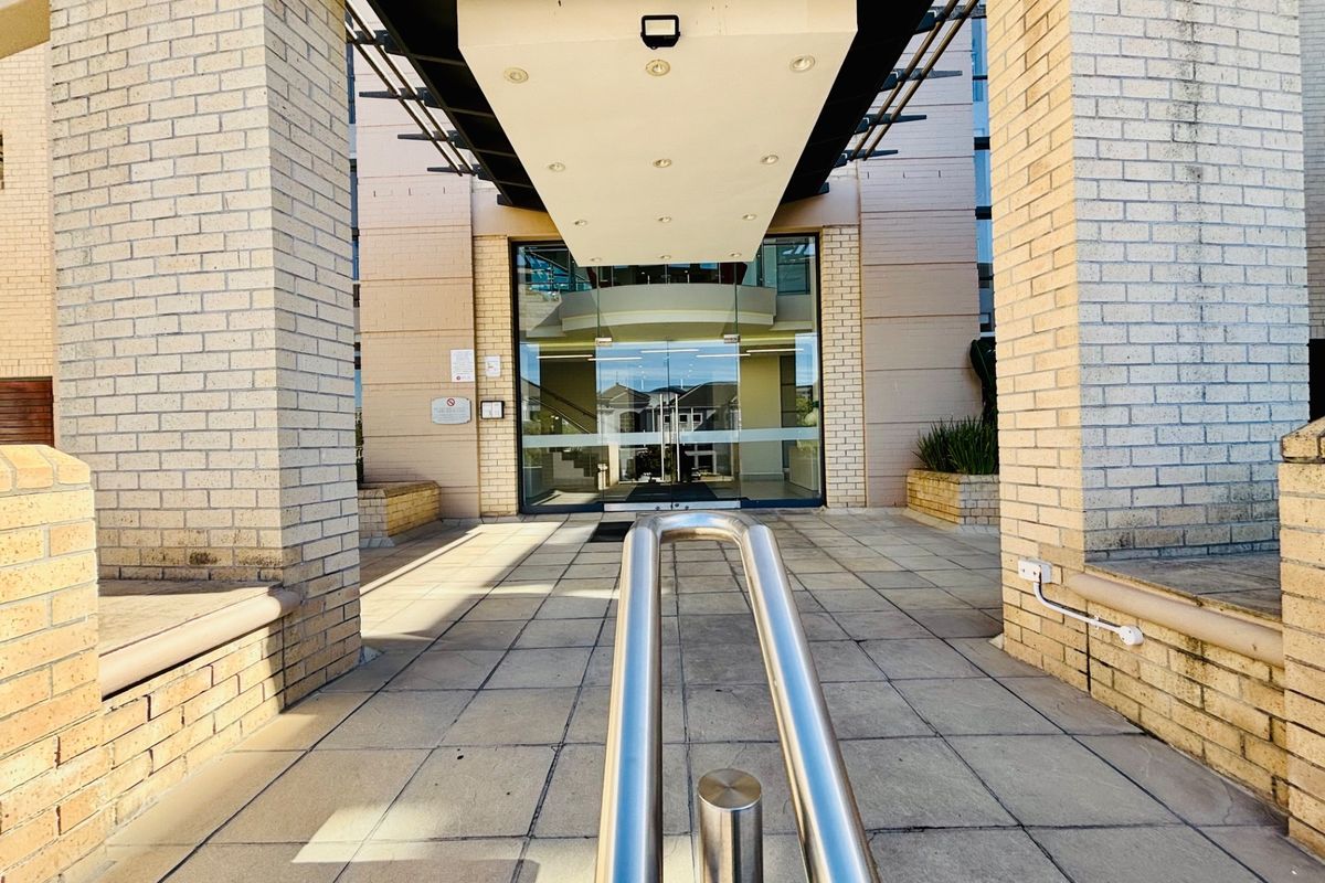 Main office building entrance featuring a paved walkway, stainless steel handrail, and a covered canopy leading to glass double doors.