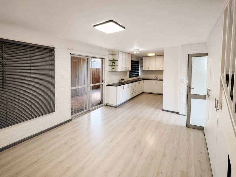 A bright, open-plan living and kitchen area featuring modern white cabinetry, laminate flooring, and a sliding door leading to an outdoor patio space.