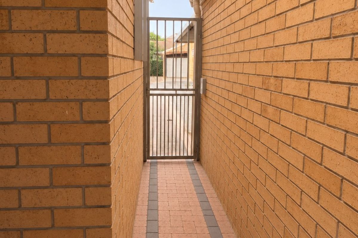 A narrow, paved side passage featuring brick walls, a secure metal gate, and a protective overhead canopy.