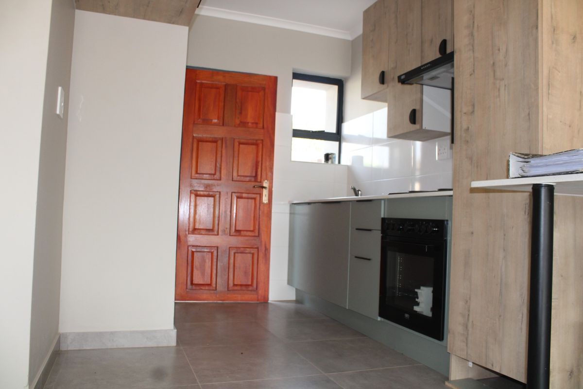 A compact kitchen featuring modern grey cabinetry, built-in oven, extractor fan, tiled flooring, and a solid wood door leading to the exterior.