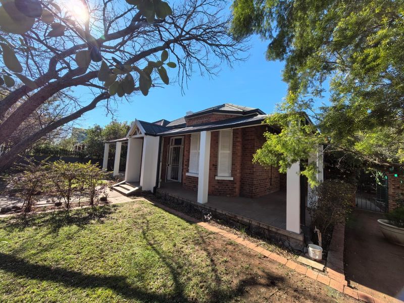 A charming brick house featuring a covered front veranda with concrete columns, set within a landscaped garden with mature trees.
