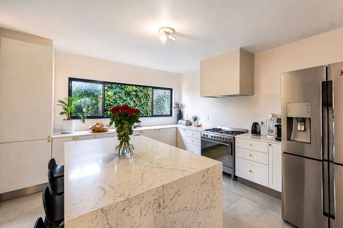Modern kitchen featuring a large stone-topped island, gas stove with extractor, ample built-in cabinetry, and large windows overlooking a lush garden.