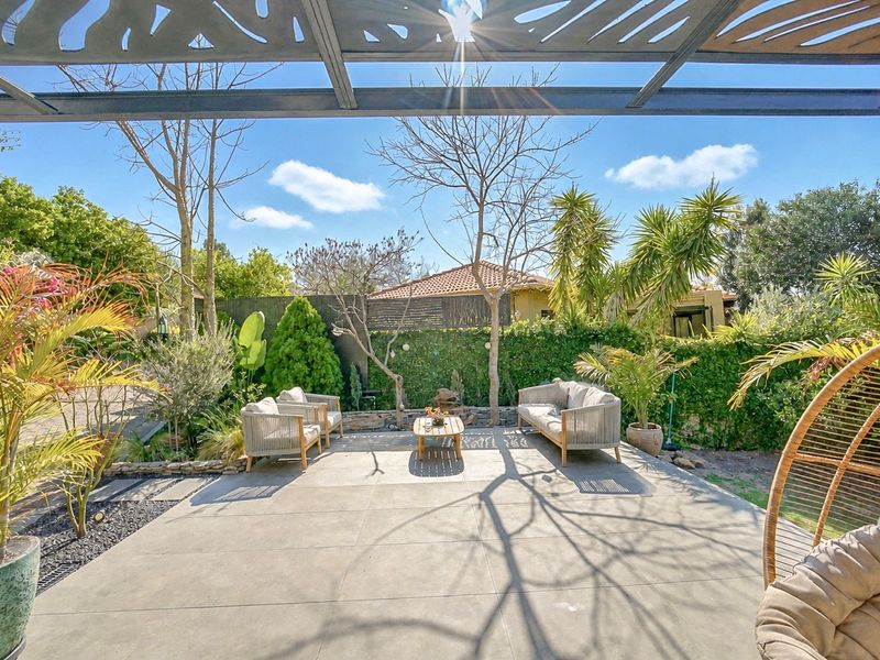 A well-maintained outdoor patio area featuring large format floor tiles, a modern metal pergola, and a lush, landscaped garden backdrop.