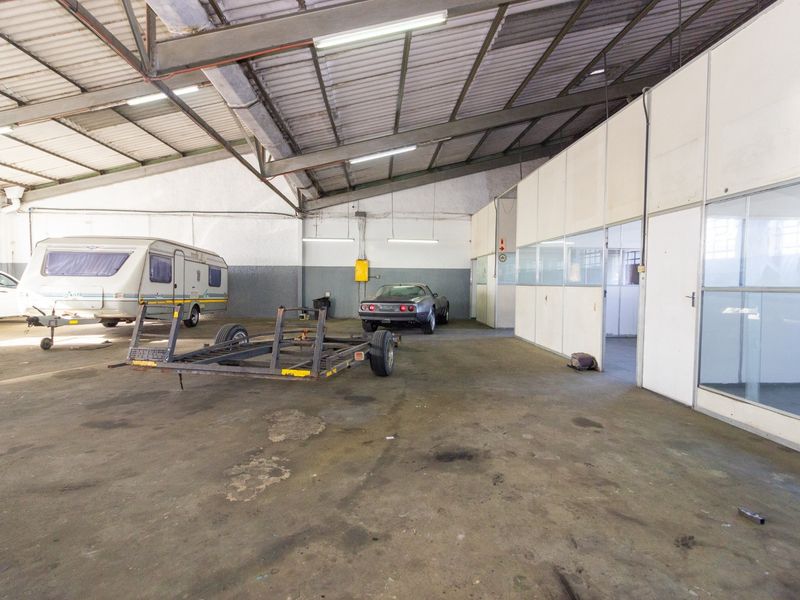 Spacious industrial garage featuring exposed steel roof trusses, fluorescent lighting, and partitioned office space with glass doors, suitable for storage or workshop use.