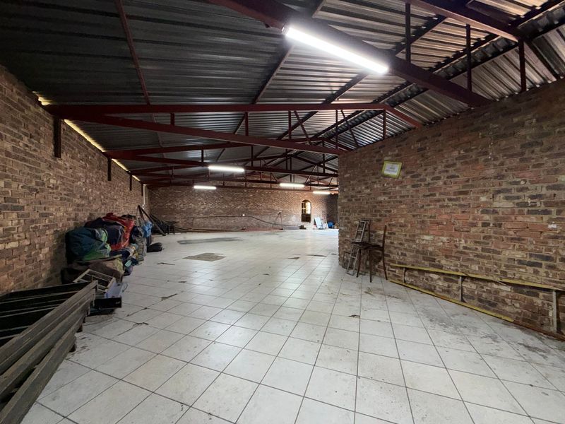 Spacious industrial utility area featuring exposed brick walls, steel roof trusses, and tiled flooring, suitable for storage or workshop use.