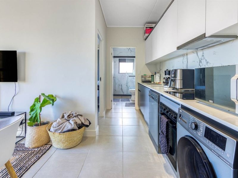 Modern open-plan kitchen featuring sleek cabinetry, stone countertops, marble-look backsplash, and integrated appliances, leading to a tiled hallway and bathroom.