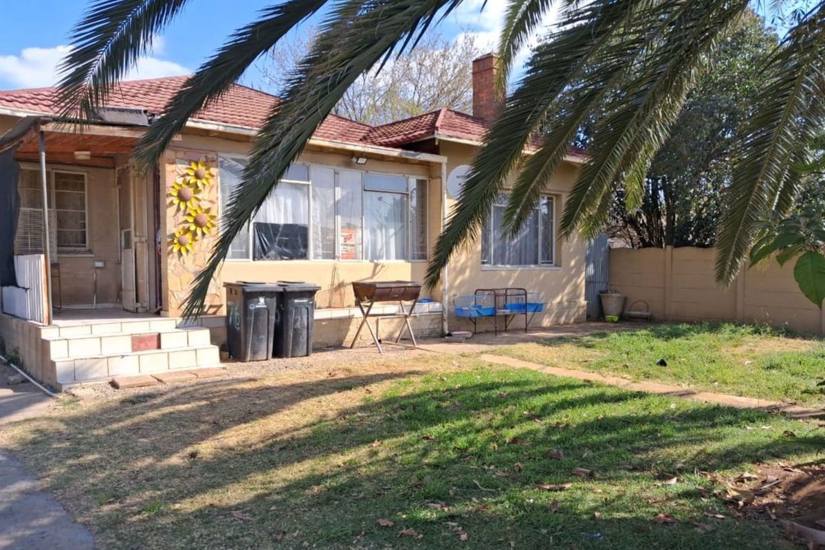 Rear view of a single-story house featuring a tiled roof, brick chimney, and a concrete patio area overlooking a garden with boundary walling.
