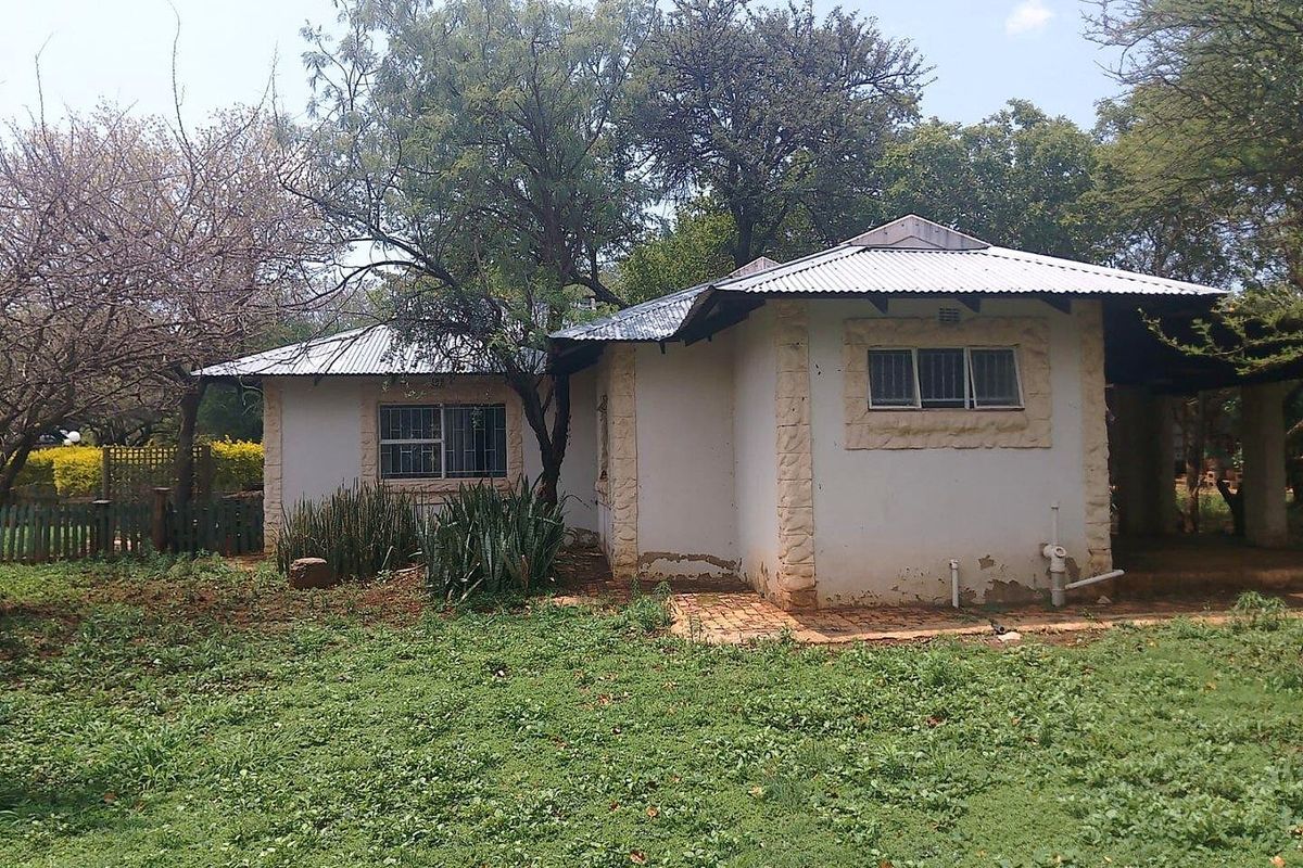 Rear view of a small holding dwelling featuring a corrugated metal roof, stone corner detailing, and security bars on windows, set within a large garden.