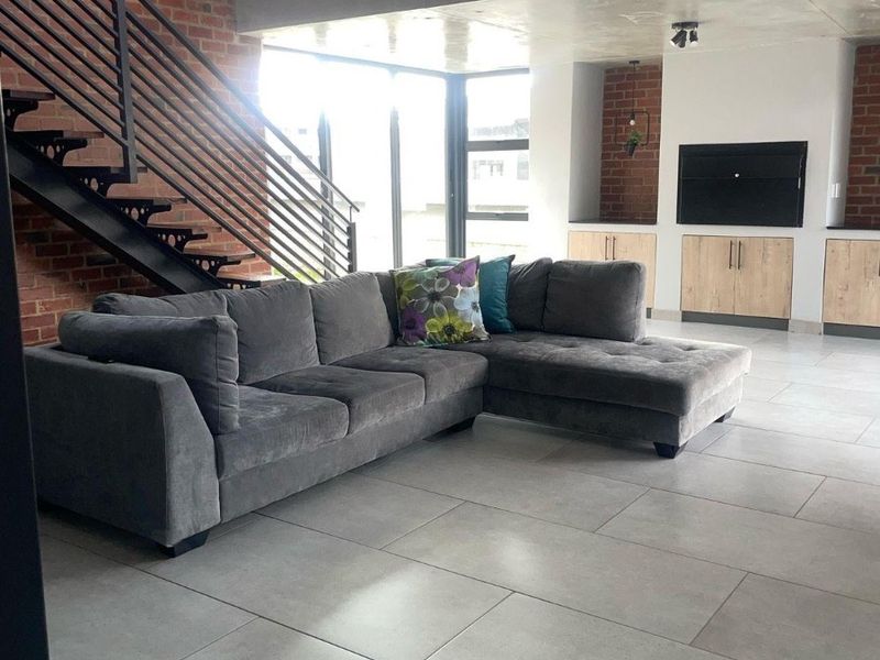 Modern open-plan living area featuring exposed brick walls, a sleek metal staircase, concrete ceiling details, and large windows providing ample natural light.