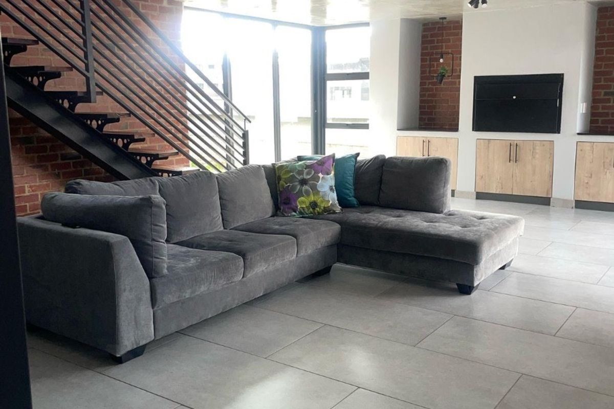 Modern open-plan living area featuring exposed brick walls, a sleek metal staircase, concrete ceiling details, and large windows providing ample natural light.