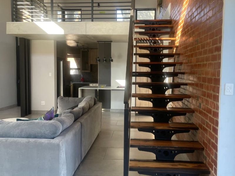 Modern open-plan living area featuring an industrial-style timber and metal staircase, exposed brick wall, and a mezzanine level overlooking the kitchen.