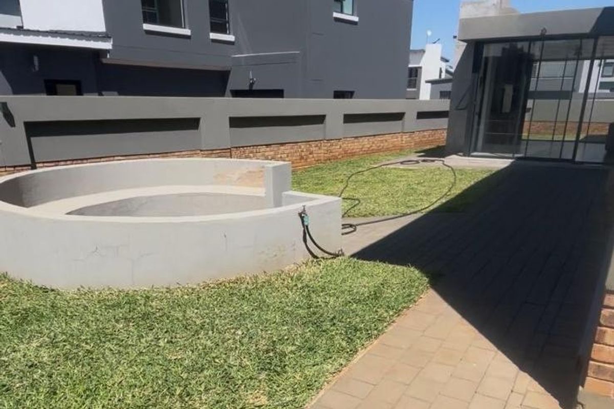 A well-maintained rear garden featuring a paved walkway, a circular built-in fire pit, and a modern two-story residential structure with a boundary wall.