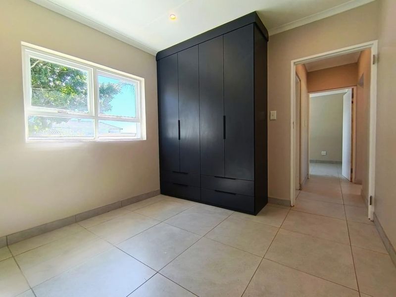 A bright, empty bedroom featuring neutral tiled flooring, a modern dark built-in wardrobe, and a large window providing natural light.