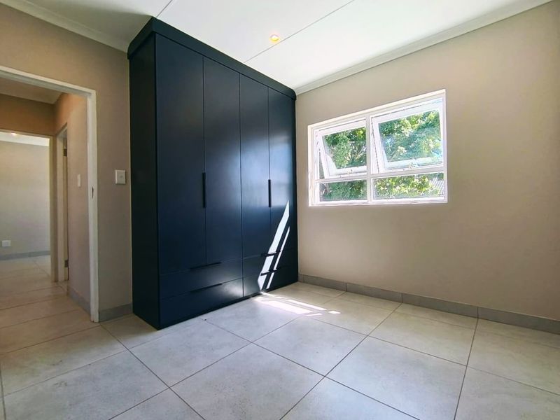 A bright, empty bedroom featuring modern dark built-in cupboards, neutral tiled flooring, and a large casement window allowing for natural light.