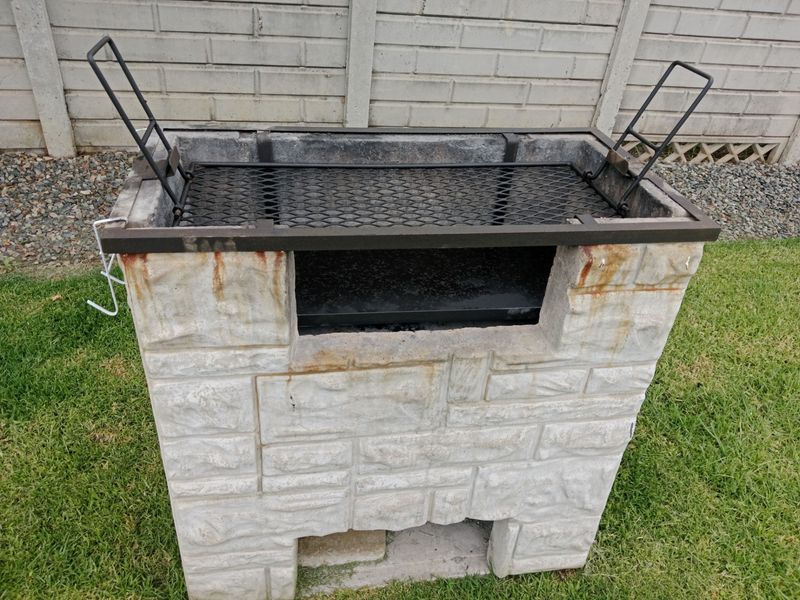 A standalone stone-built outdoor braai area featuring a metal grill grate, situated in a garden setting.