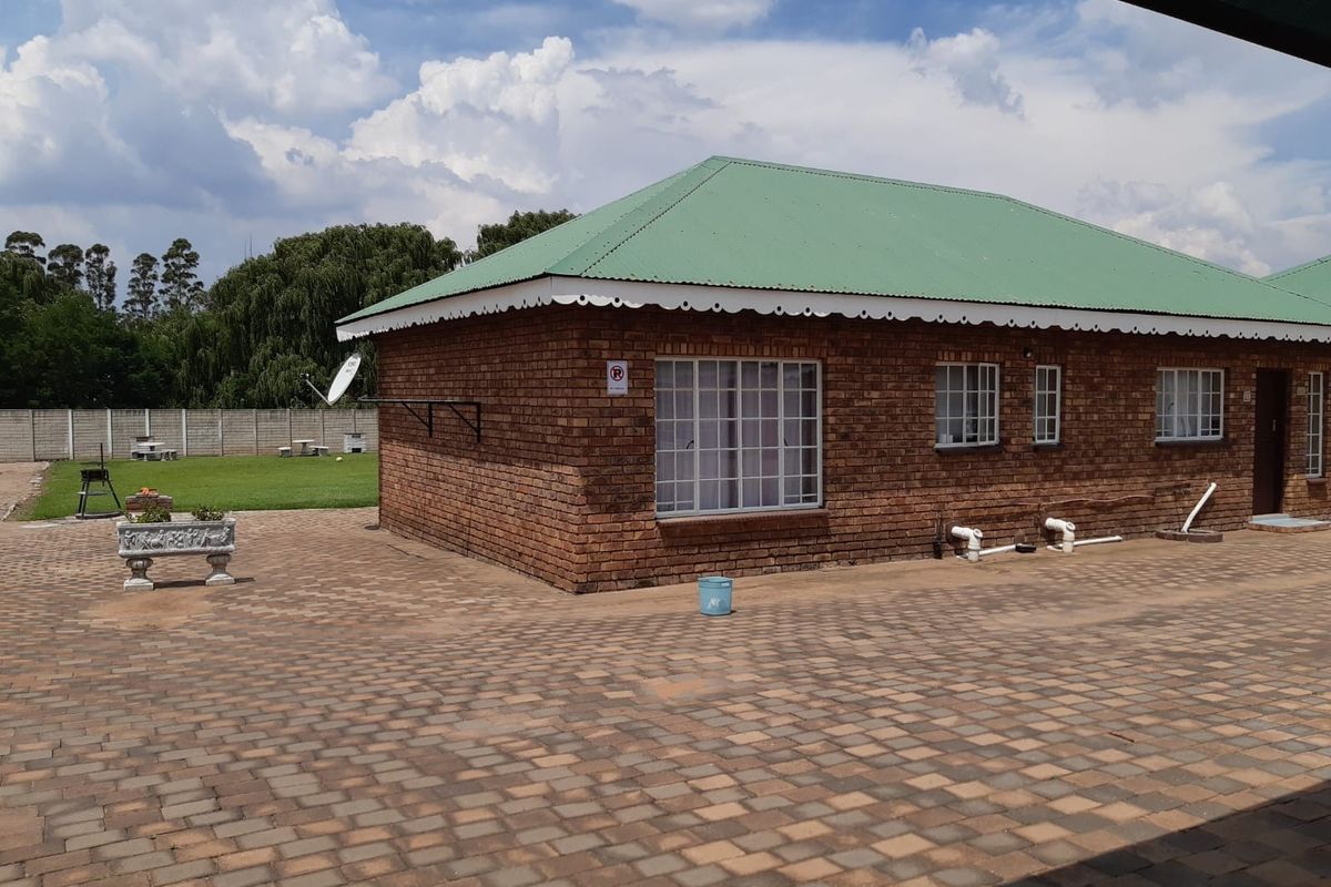 A rear view of a brick house featuring a green metal roof, paved patio area, and a surrounding perimeter wall.