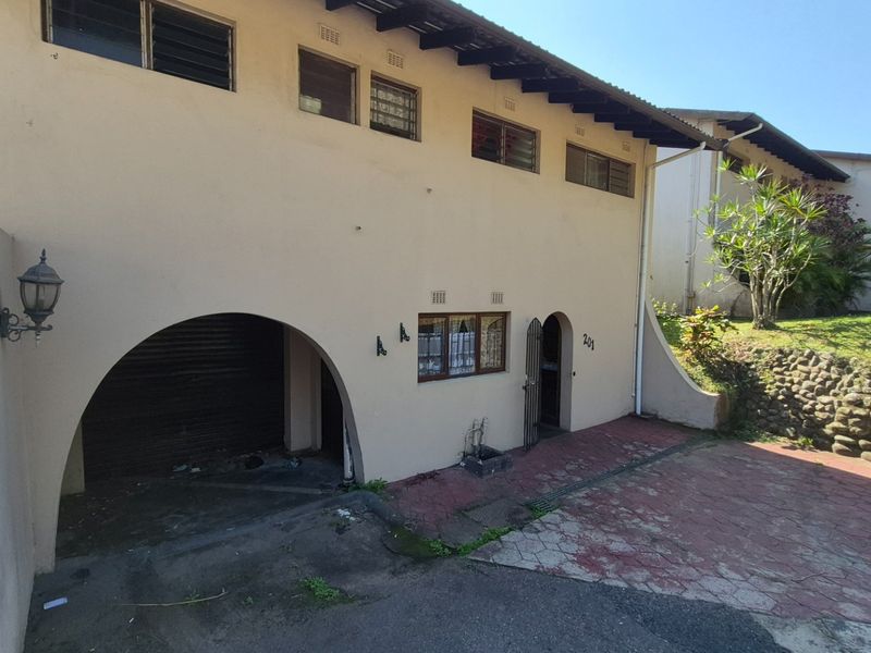 Front elevation of the property featuring an arched garage entrance, paved driveway, and louvered windows with visible exterior wear.
