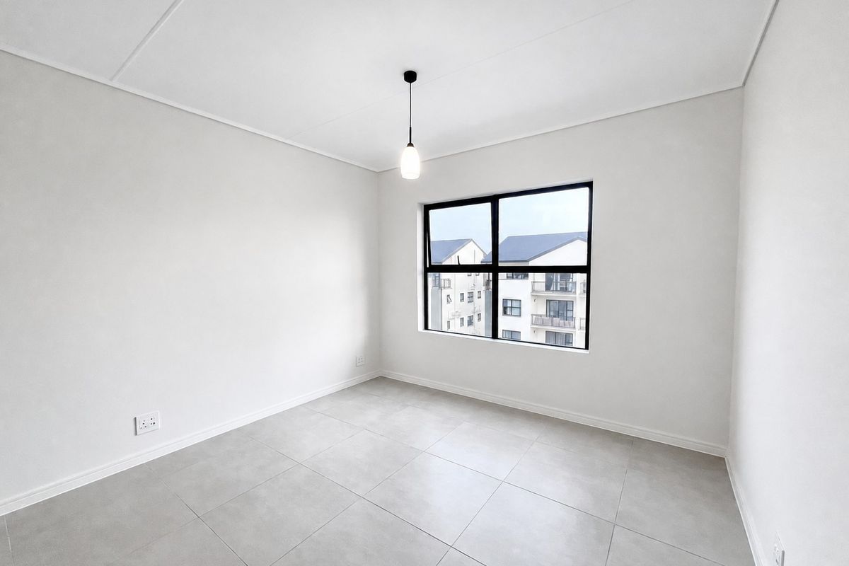 A bright, empty bedroom featuring neutral tiled flooring, a modern pendant light, and a large black-framed window with views of the surrounding apartment complex.