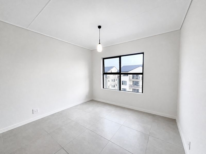 A bright, empty bedroom featuring neutral tiled flooring, a modern pendant light, and a large black-framed window with views of the surrounding apartment complex.