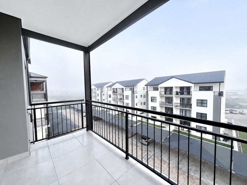 A clean, modern covered balcony featuring tiled flooring and a black metal balustrade, offering an elevated view of the surrounding apartment complex.