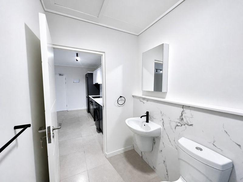 Modern bathroom featuring a wall-mounted basin with black fixtures, marble-look tiling, and a toilet, with an adjacent view into the kitchen area.