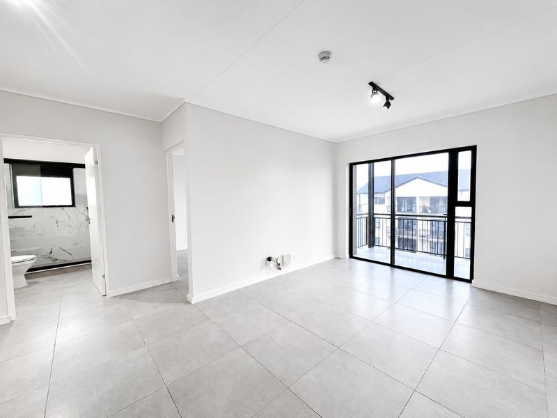 A bright, open-plan living area featuring modern tiled flooring, track lighting, and a large sliding glass door leading to a balcony.