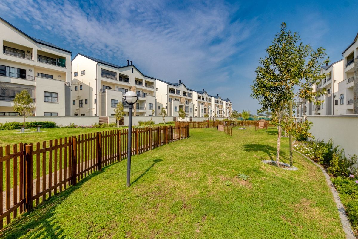 A well-maintained communal garden area featuring manicured lawns, perimeter fencing, and paved walkways set against a modern apartment complex.