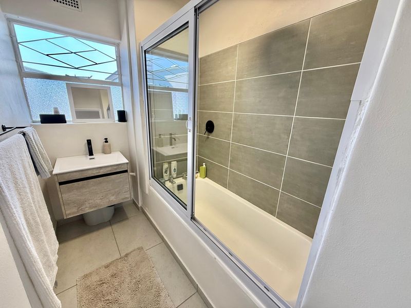 Modern bathroom featuring a vanity with basin, a bathtub with a sliding glass screen, and neutral wall and floor tiling.