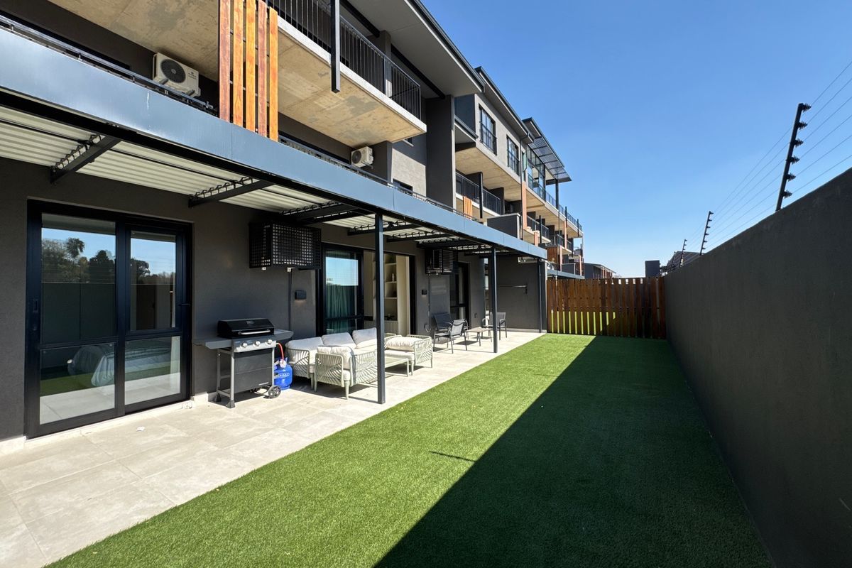 A modern ground-floor apartment exterior featuring a covered patio, artificial lawn, and secure perimeter wall with electric fencing.