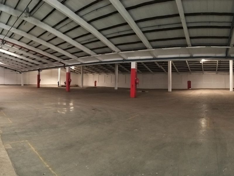 Expansive industrial warehouse space featuring high ceilings with exposed steel trusses, concrete flooring, and a mezzanine access staircase, suitable for storage or manufacturing.