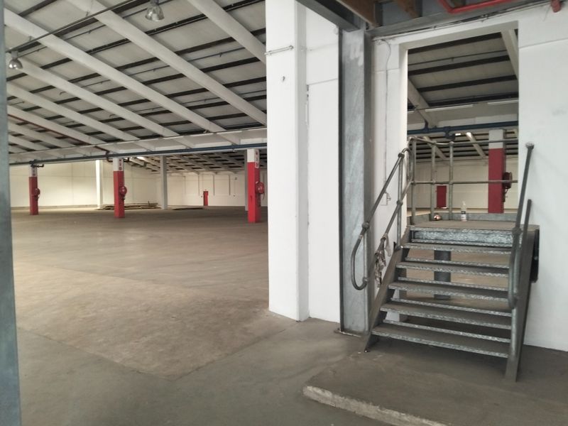 Spacious industrial warehouse area featuring exposed steel roof trusses, concrete flooring, and a metal staircase leading to an elevated section.