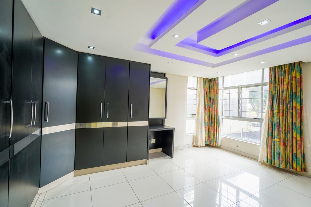 Spacious bedroom featuring extensive built-in cupboards, a dedicated vanity area, modern recessed ceiling lighting with accent features, and large windows with security bars.