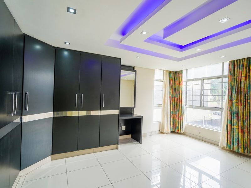 Spacious bedroom featuring extensive built-in cupboards, a dedicated vanity area, modern recessed ceiling lighting with accent features, and large windows with security bars.