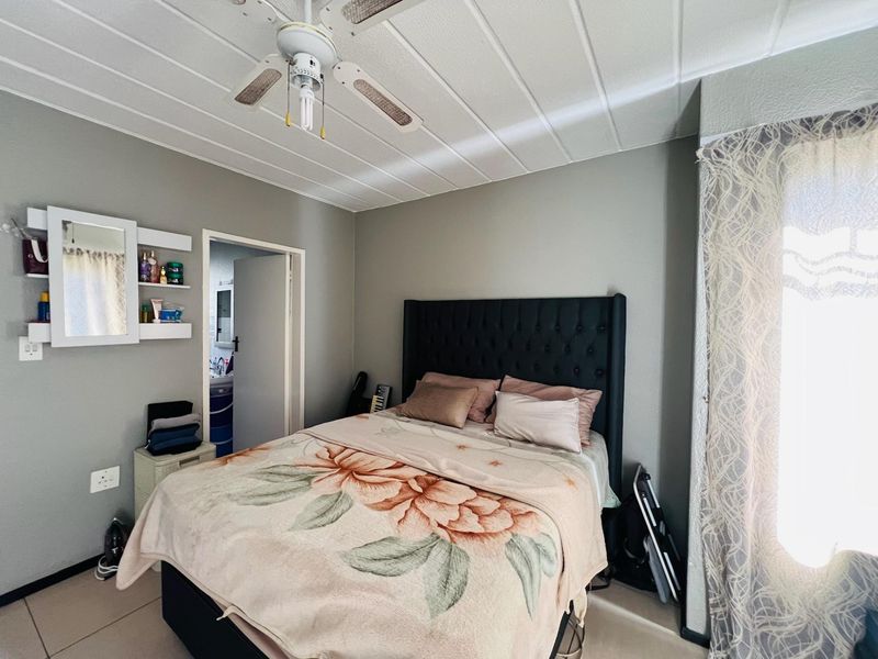 A bedroom featuring tiled flooring, a panelled ceiling, a ceiling fan, and built-in wall shelving, with an adjoining doorway leading to an en-suite bathroom.