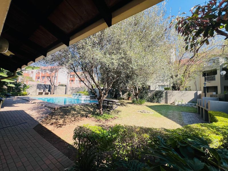 A well-maintained communal outdoor area featuring a swimming pool, paved patio, built-in braai facilities, and established garden landscaping.