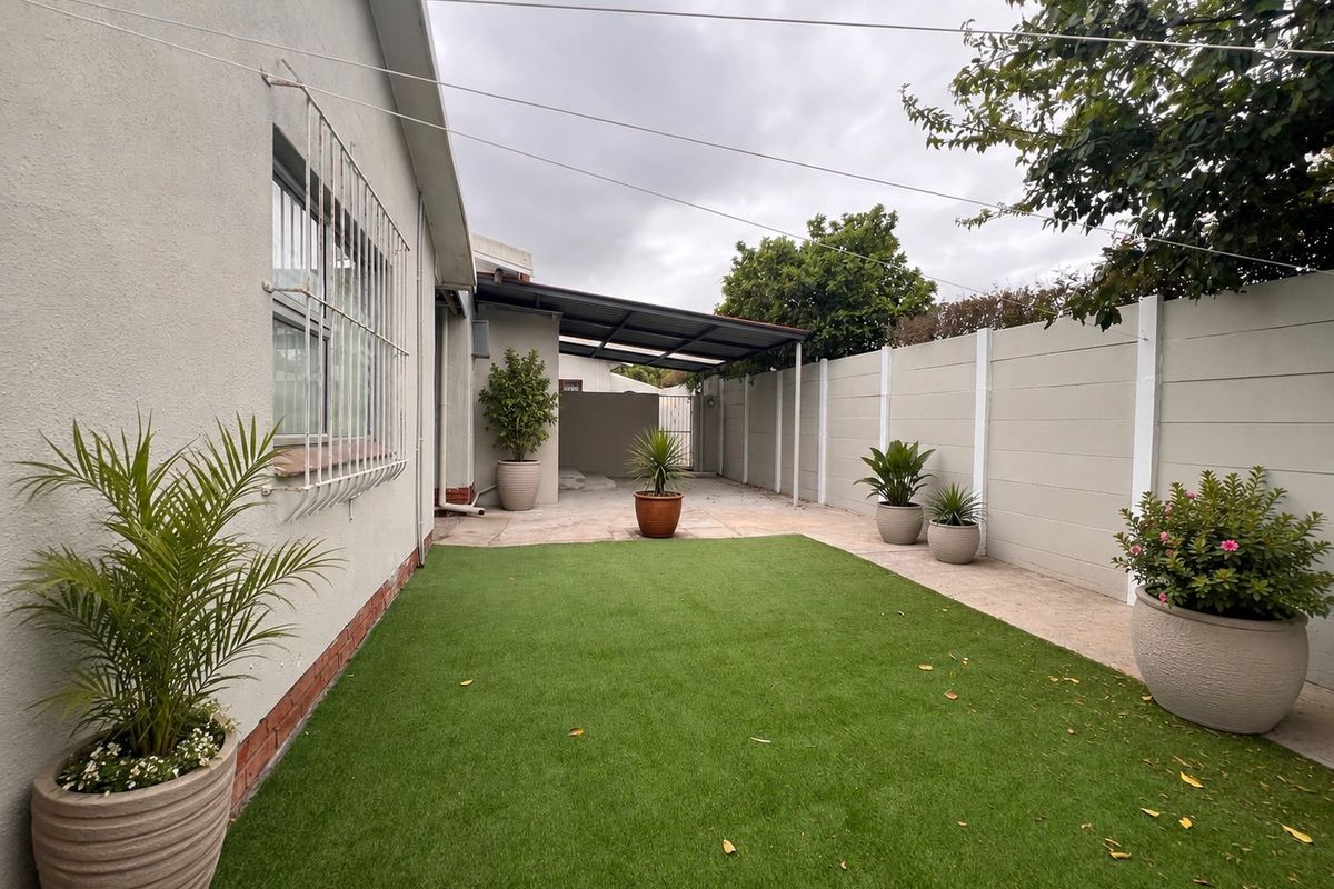 A neat rear exterior space featuring artificial grass, a paved patio area, and a covered section enclosed by a precast concrete boundary wall.