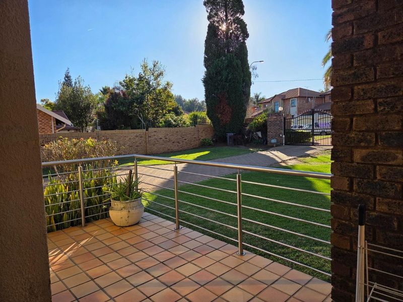 A covered patio area featuring tiled flooring and a modern stainless steel balustrade, overlooking a private garden and paved driveway.