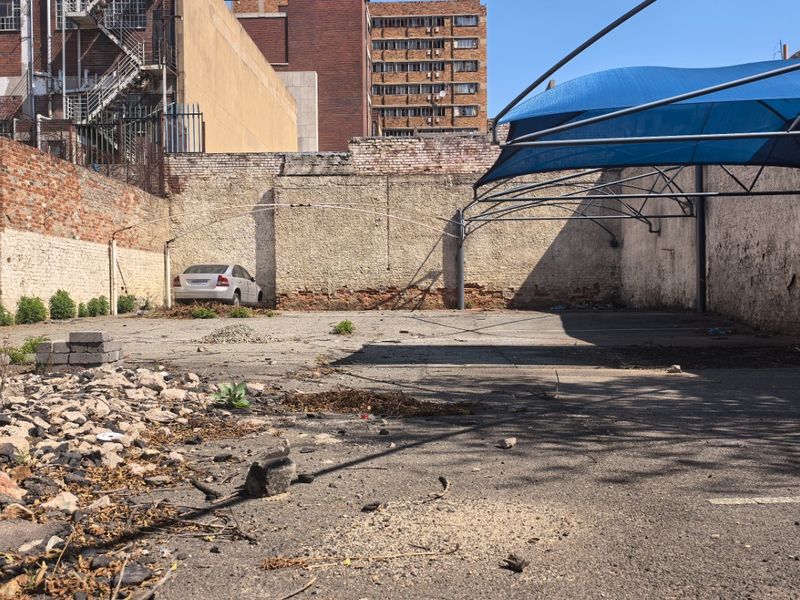 A vacant commercial paved lot featuring a shade sail structure and perimeter brick walls, showing significant signs of wear and surface deterioration.