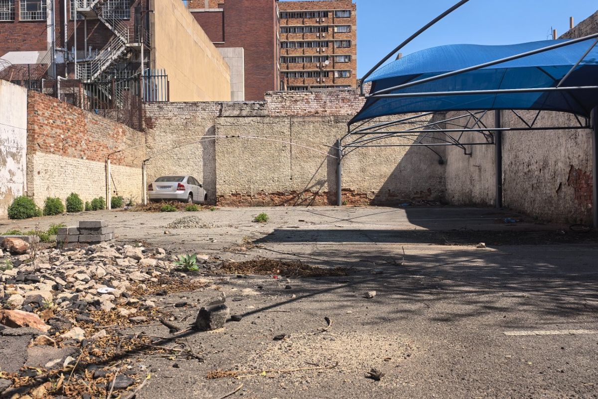 A vacant commercial paved lot featuring a shade sail structure and perimeter brick walls, showing significant signs of wear and surface deterioration.