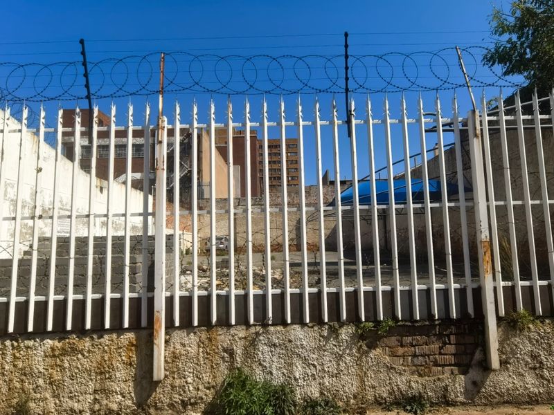 Street-facing view of a vacant commercial plot secured by a white steel palisade fence with razor wire and a weathered retaining wall.
