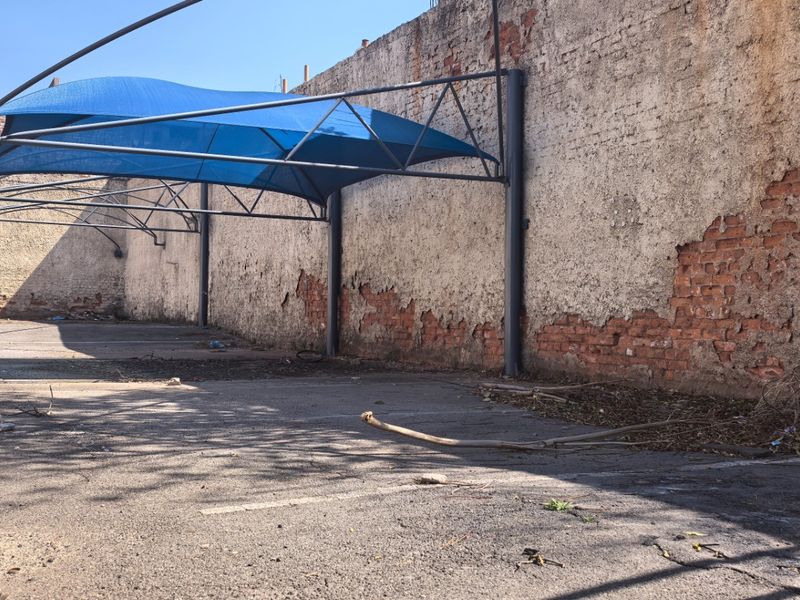 Vacant commercial land featuring an asphalt parking area with a blue shade cloth carport structure and a perimeter brick wall requiring maintenance.